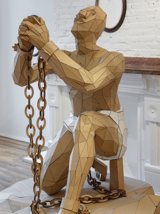 Sculpture. A man made of mostly triangular pieces of cardboard kneels on one knee. His other foot is forwards, his hands clasped and raised. Chains connect shackles on his wrists and knees. His head tips back. His body is angled to our left in this view.