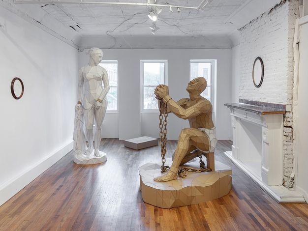 Installation. Two sculptures are made with triangular and trapezoidal pieces of cardboard. One shows a kneeling man with chains hanging from manacles on his wrists and ankles. His skin is the colour of the brown cardboard. The other sculpture shows a nude woman standing and looking over one shoulder, her wrists chained together. That whole sculpture is white except for the manacles.