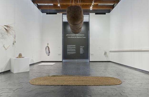 Installation. A long wooden object hangs from the ceiling over a woven mat on a grey stone floor in a gallery with white walls. More sculptural pieces including a bell and handmade brooms hang along the side walls or in the far corner.