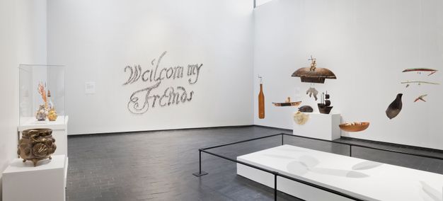 Installation. From one corner we look across a room with a grey stone floor and white walls. Sculptures sit on rectangular white bases, and other works, perhaps mobiles, hang from the ceiling in a group over a low white platform. Writing on the far wall appears to be made with tiny brown dots, and it reads, “Weilcom my Friends”.