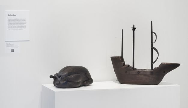 Installation. Two darkened wood sculptures sit on a white base. One appears to be a plucked and decapitated turkey, either roasted or read to be so. The other is a three-masted ship.