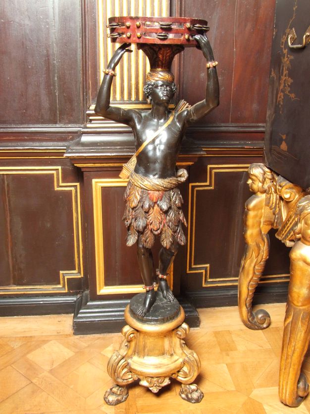 Sculpture. Carved from wood and painted, a man with dark brown-coloured skin stands on an ornamental base and balances on oversized tambourine on his head. A tapering foot under the tambourine sits on a gold turban, and the man wears a skirt of leaves or feathers.