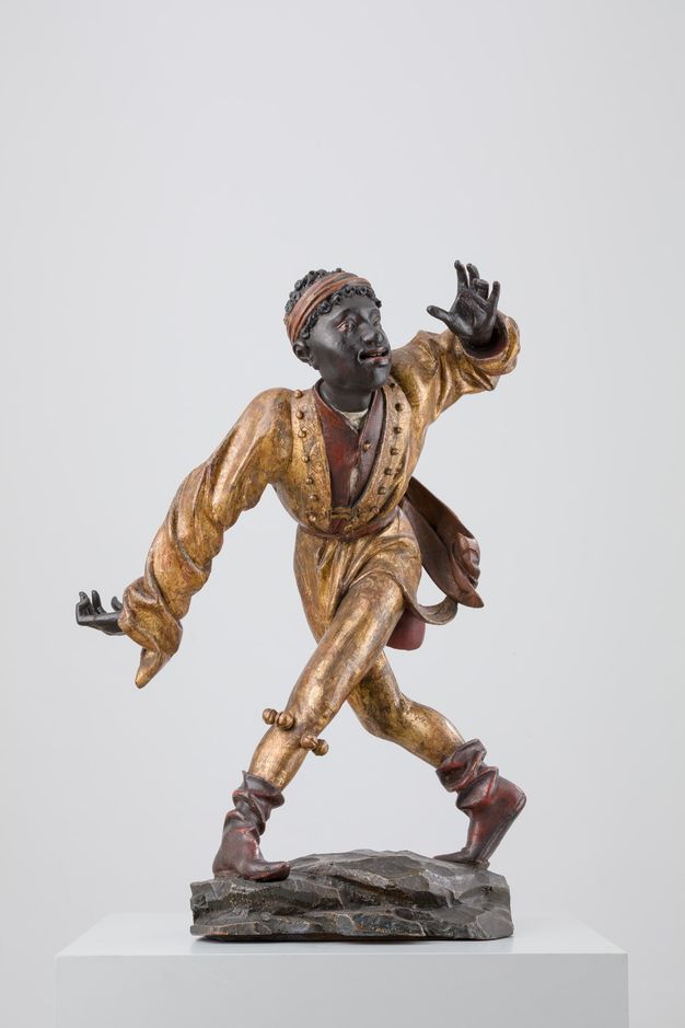 Sculpture. Carved from wood and then painted, a man with dark brown skin is shown mid-dance step. One leg crosses far in front of the other, feet planted wide. An extended arm matches the angle of the leg, and the other hand is raised. The person’s red lips are parted, and they look up and to our right.