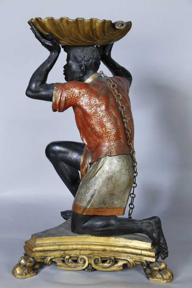Sculpture. Carved from wood and then painted, a man with dark brown or black skin kneels on a triangular gold base. The man faces our left in this view, so we see a gold chain hanging down his back from a gold band around his neck. He balances a gold shell-shaped dish on his head.