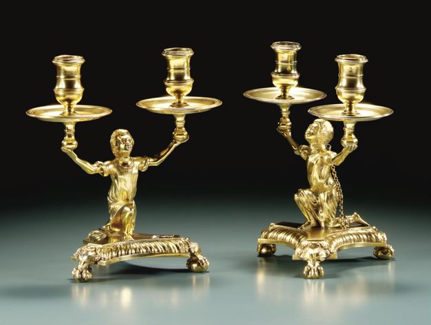 Metalwork. Two gold candelabra are in the shape of kneeling men each holding up a pair of capitals above drip pans. One of the candelabra to show a chain connecting a shackle around one man’s neck to an ankle.