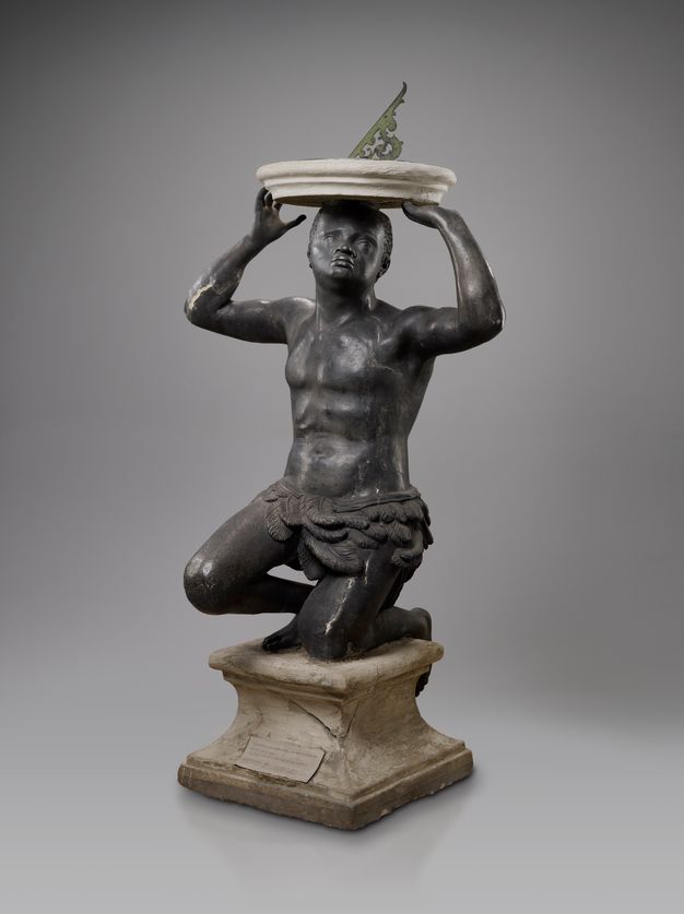Sculpture. A man cast in in bronze balances a disc with a sundial on his head as he kneels on a pedestal base. The man is nude except for leaves or feathers around his waist.