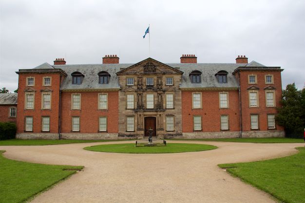 Architecture. A three-storey brick and stone mansion with four wide chimneys is at the end of a circular drive. At the centre of the drive, a statue of a person holding up a sundial can just barely be made out.