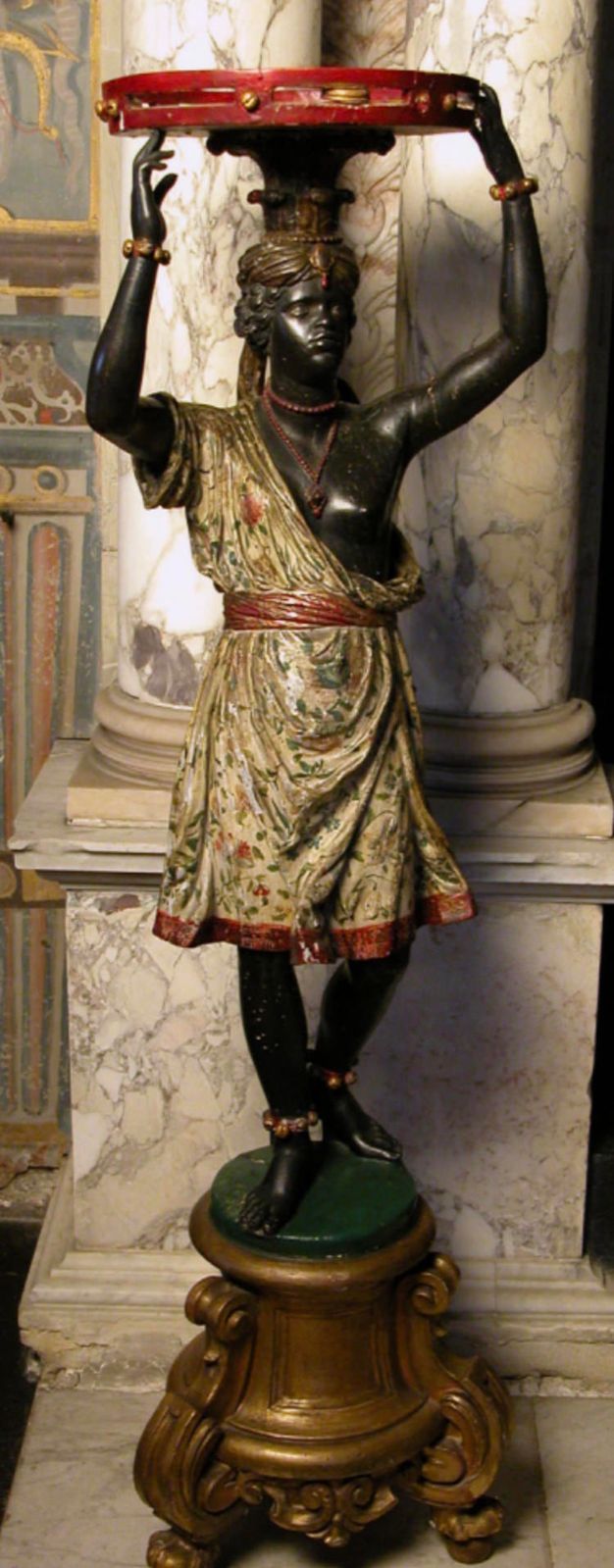 Sculpture. Carved from wood and then painted, a woman with black-coloured, polished skin stands on an ornate base and holds a wide, red tambourine balanced on her head. Her floral-patterned toga falls off one shoulder to reveal a breast. Her wrists and ankles are encircled with gold.