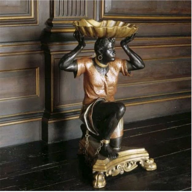 Sculpture. Carved from wood and then painted, a man with dark brown or black skin kneels on a triangular gold base, a gold chain connecting ankle shackles. He balances a gold shell-shaped dish on his head. Another gold band is around his neck, and he wears a gold hoop in the earring we can see.