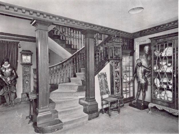 Black and white photograph. A zig-zagging staircase framed by fluted piers creates a corner, in which stand a chair, grandfather clock, two glass-fronted cabinets full of ceramic, and a life-sized, free-standing person. Details are difficult to make out in this image, but the person wears a coat with tails, knee-length breeches and stockings, and a long vest.