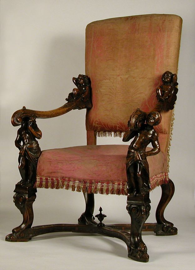 Chair. The back and seat are upholstered with a faded red, floral brocade fabric. Polished wood arms sweep forwards and are supported by carved people who create the supports. The pedestals the people stand on become the front legs. Each person sways outwards like backwards parentheses.