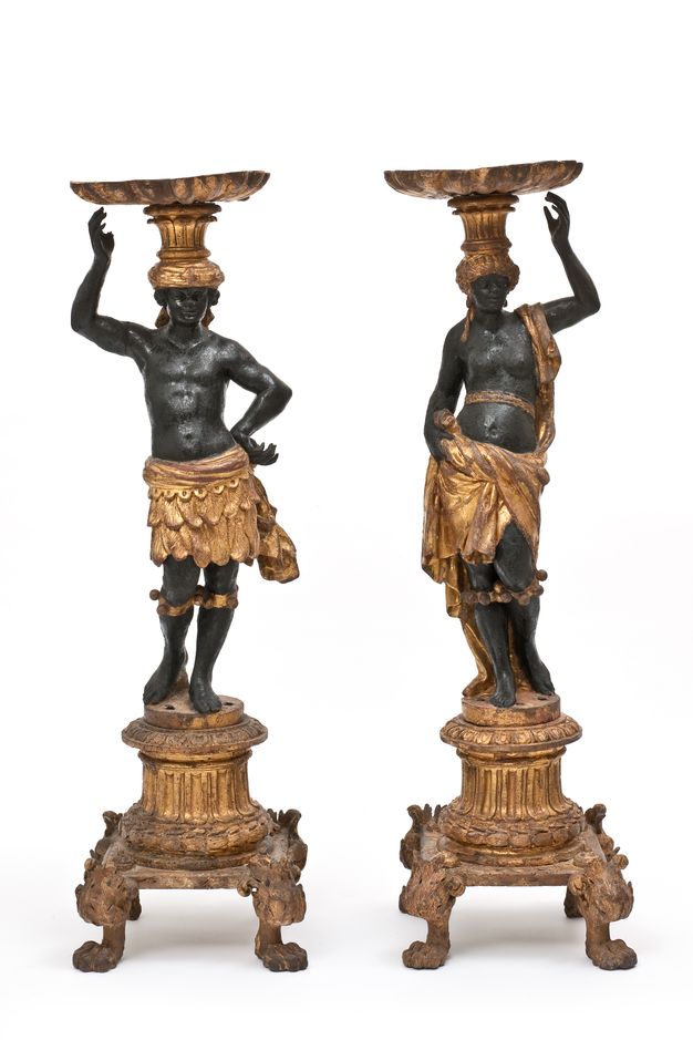 Sculptures. Carved from wood and then painted, a man and woman with black-coloured skin stand on gold pedestal bases and balance gold dishes on their heads. They are both bare-chested with fabric or a skirt around their hips. Bands with bells are around their legs just under their knees.
