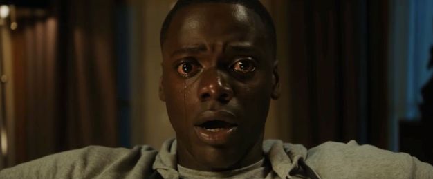 Video still. Close-up view of a dark-skinned man with eyes wide open, tears streaming down his cheeks. His lips are parted.
