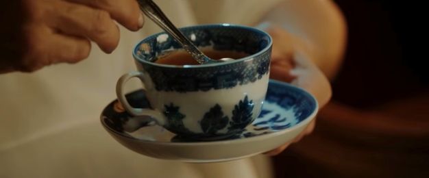 Video still. Close-up view of a blue and white porcelain teacup. A pale-skinned hand holds a silver spoon in the brown tea.