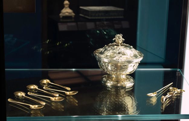 Installation. A covered dish with a flaring foot is surrounded by a tea strainer, sugar cube tongs, and six teaspoons. The objects reflect in the glass at the back of the case and in the glass shelf on which they sit.