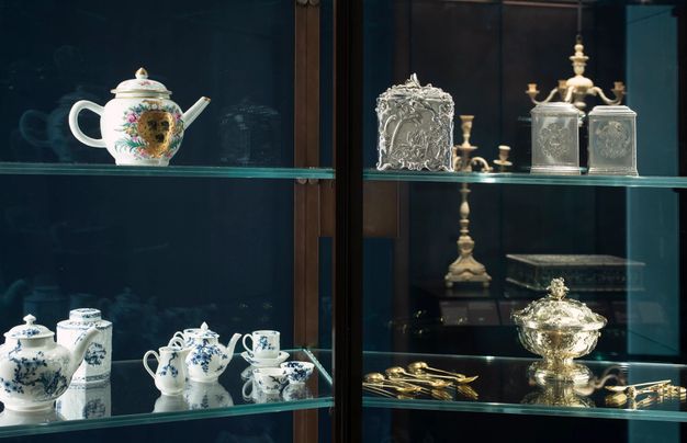 Installation. Four glass shelves in a cabinet hold porcelain, silver, and gold vessels, containers, and utensils for storing and making tea. The objects reflect in the glass on the back side and in the glass of the shelves, creating double and sometimes triple vision.
