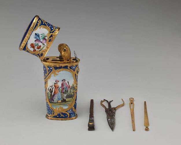 An enamelled copper container has openings for four implements that lie alongside it in this image. Two appear to be tweezers, one might be nail clippers, and the fourth is a pair of small scissors. The box is decorated with a scene of two pale-skinned people walking in a landscape.