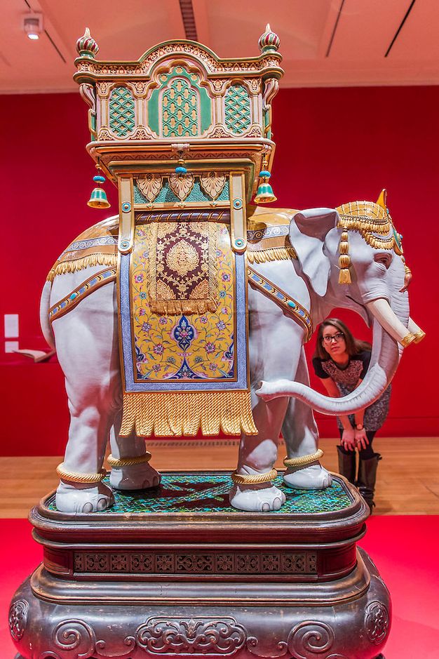 Sculpture. An elephant is draped in gold cloth patterned with blue and red below a balcony-like structure called a howdah. A woman peering through the oval space left by the elephant’s trunk gives a sense of scale. The complete earthenware sculpture is about half again as tall as the woman would be.