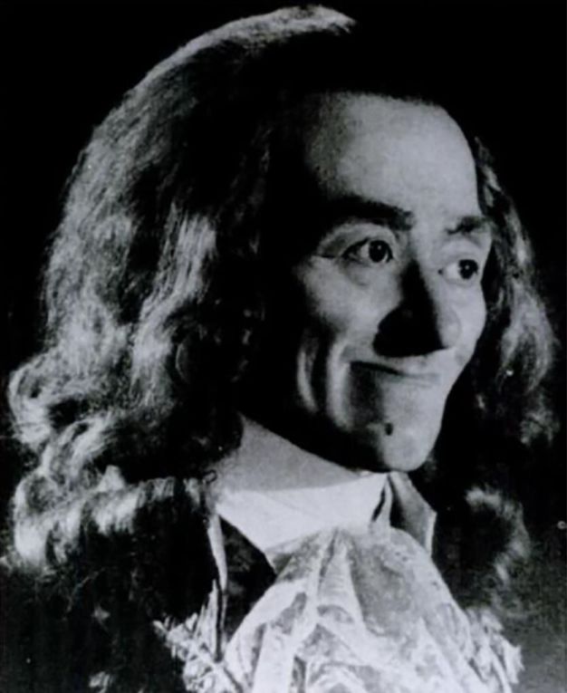 Black and white photograph. A man with thin lips pressed together in a smile looks off to our right. He has thick, dark brows, and his long hair hangs loose. He wears a high-collared white shirt and a frilly cravat.