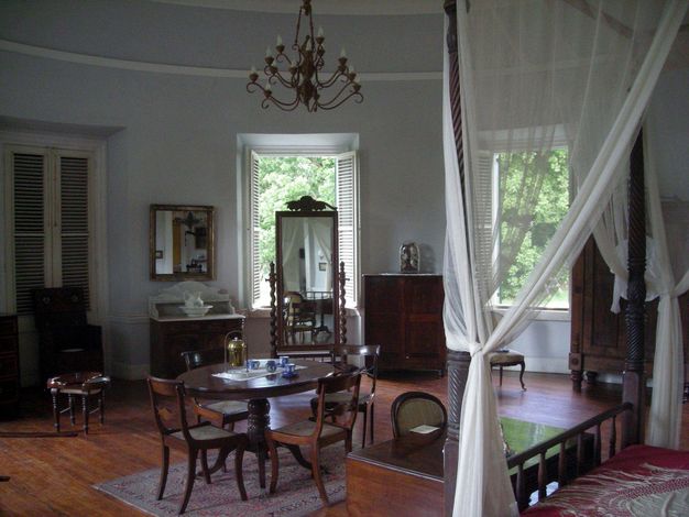 Architecture. Tables, dressers, a full-length mirror, and a canopied bed are arranged in a room with two windows that open onto trees. A two-tiered chandelier hangs above, and porcelain and brass objects are on the tables and dressers.