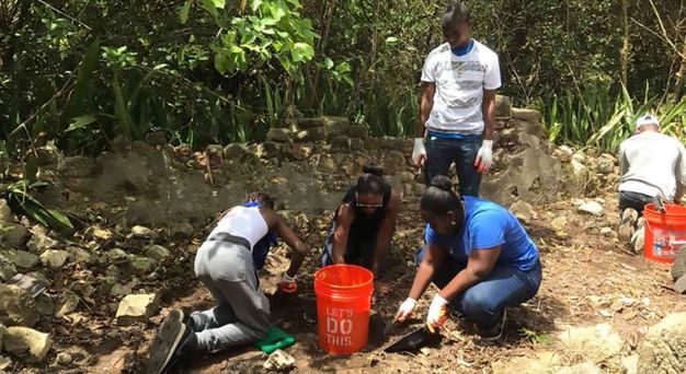 Archaeological dig. Four people work in the soil near a stone wall in dappled shade, greenery growing thickly behind them. A fifth person looks on. They all have dark skin. They wear gloves and dig or sift with spades.