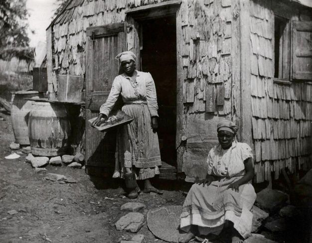 Black and white photograph. Two dark-skinned women sit and stand outside, near the opening of a door leading into a cabin. Both wear long skirts and caps.