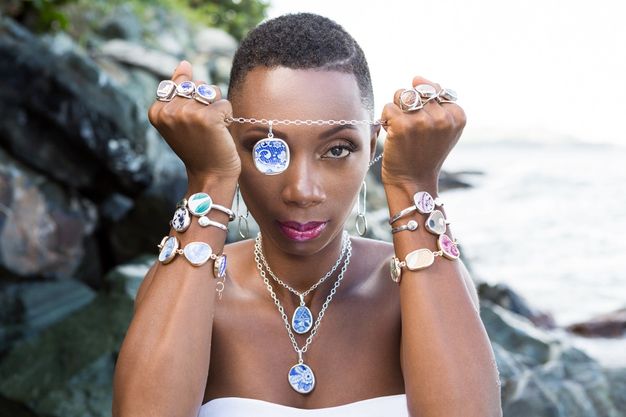 Jewellery. A dark-skinned woman wears six rings, six bracelets, and two necklaces made with fragments of blue, pink, or green-painted ceramics. She holds the chain of a third necklace up between fists at forehead level, so the blue and white pendant there overlaps one brown eye. The sea is out of focus behind her.