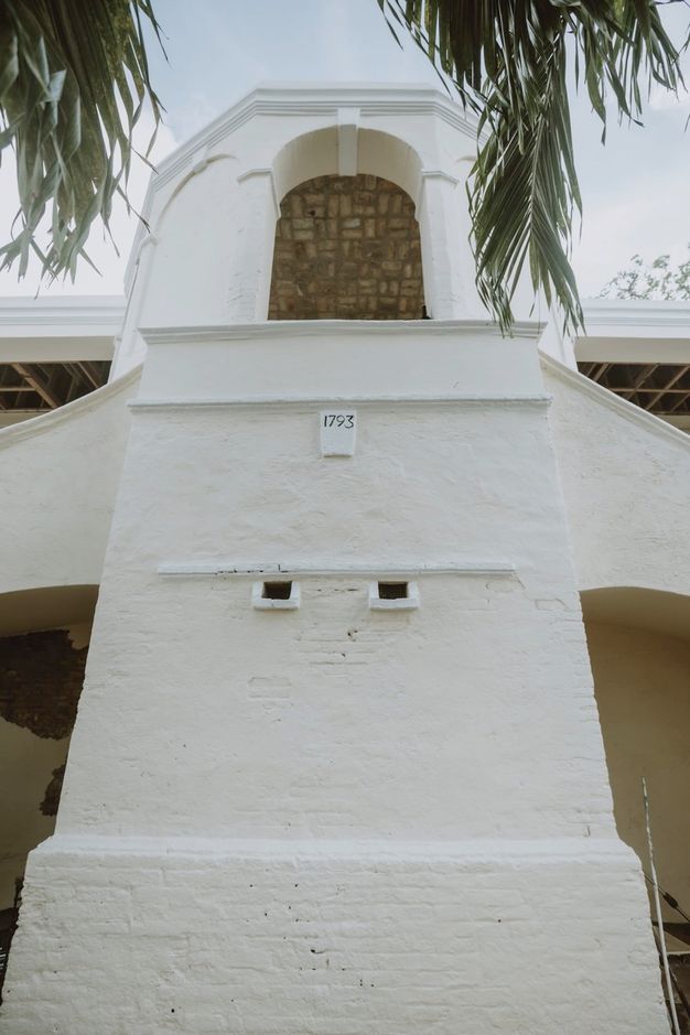 Architecture. We look sharply up at a whitewashed structure with an open arch on the second level. A plaque on the front centre of the building reads “1793”.