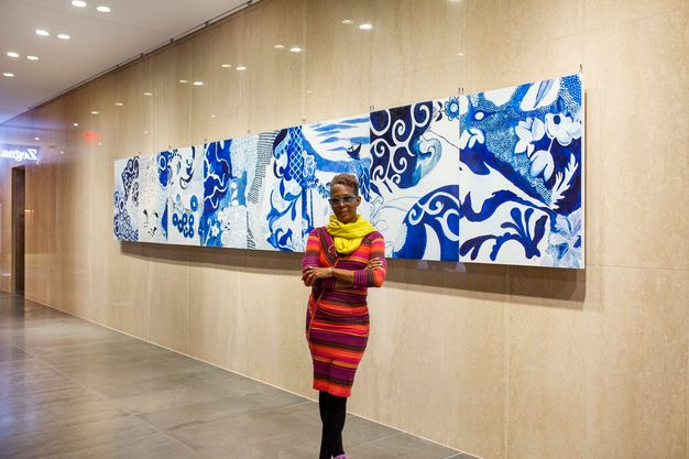Installation. A dark-skinned woman wearing a purple, orange, yellow, and green-striped dress and canary-yellow scarf stands with ankles and arms crossed in front of a long painting hung on a polished stone wall. The painting appears to have eight joined sections painted with white and shades of blue.