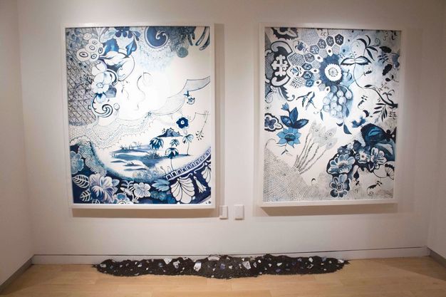 Installation. Patterns, organic forms, and landscapes painted in tones of midnight and muted blue swirl against white backgrounds in two paintings hung on a gallery wall. A long dark object lies along the floor in front of the paintings.
