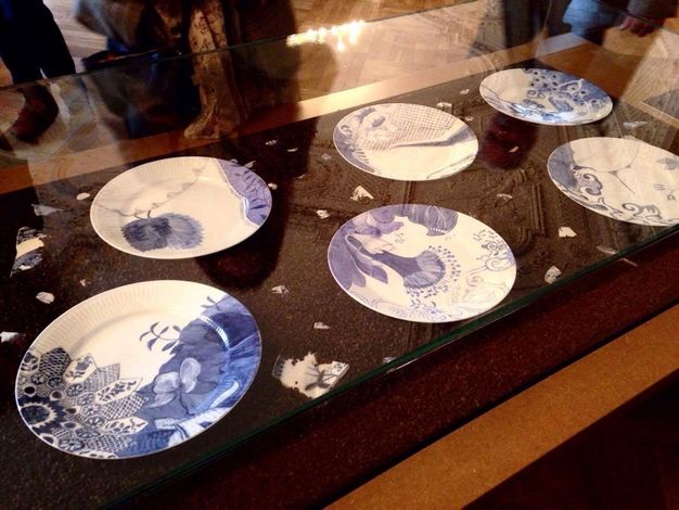 Installation. Six plates in a glass case are painted with bright, deep blue floral and geometric patterns on white porcelain. The rims of the plates are ridged. Fragments of more white porcelain painted with blue is scattered around the plates.