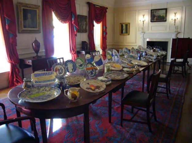 Installation. Plates and dishes are set and displayed on a long dining room table. Pale yellow panelling covers the walls and heavy scarlet-red drapes hang from the windows. Details on the plates are difficult to make out but many are colourful and are different sizes.