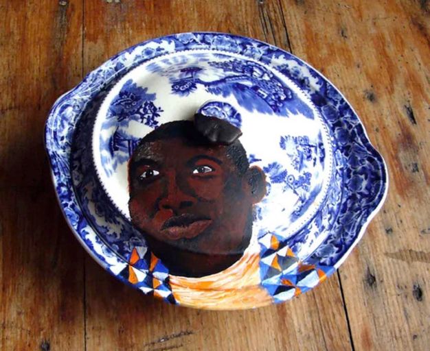 Installation. A dark-skinned man’s portrait is painted on a lidded white and blue porcelain dish. The person’s face takes up the bottom left quadrant of the dish and spans the different levels of the handle, lid, and rim.