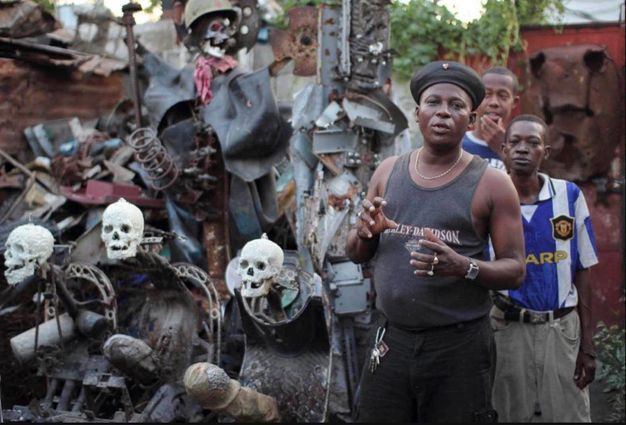Installation. Three dark-skinned men stand next to a sculptural, multimedia assemblage, which includes skulls and pieces of metal. The work appears to be as tall as the men and would take a few strides to walk across.