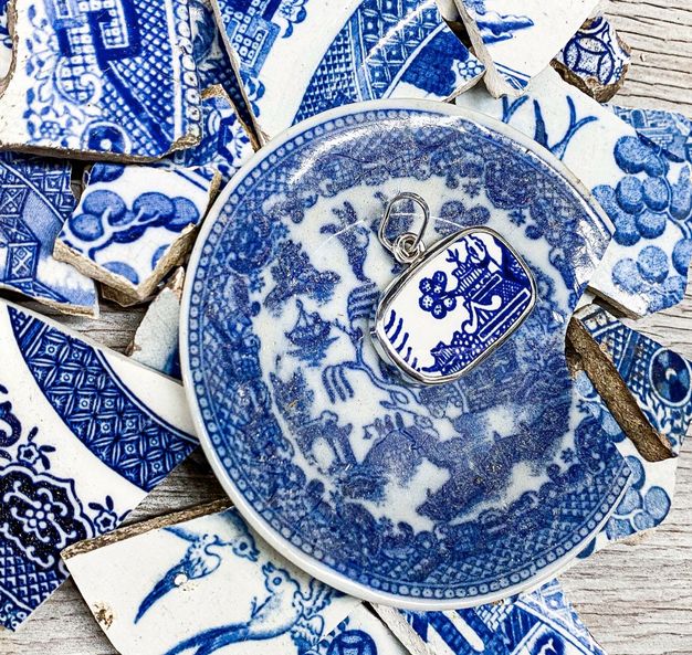 Jewellery. A piece of white porcelain painted in blue with a pagoda is made into a pendant. The pendant sits on a pile of ceramic fragments, all painted with blue against white.