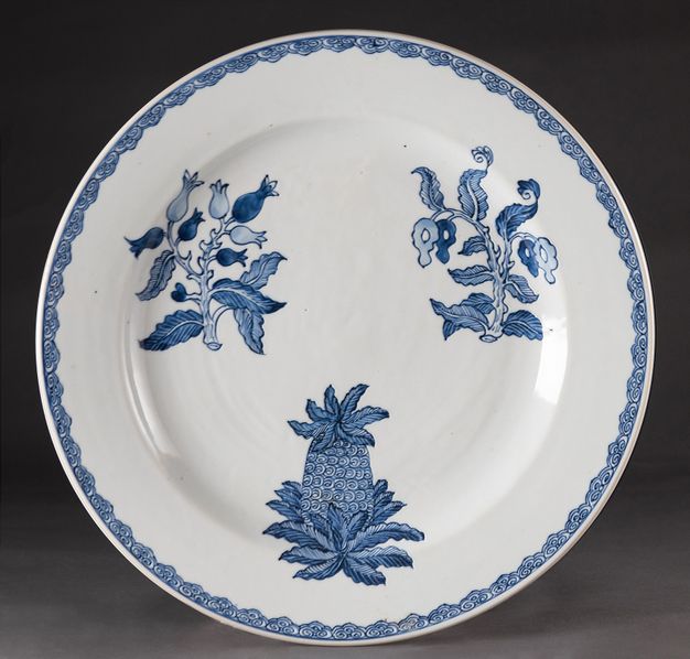 Porcelain. A white plate is painted with two specimens of flowers and one pineapple. The edge has a narrow, swirling geometric pattern.