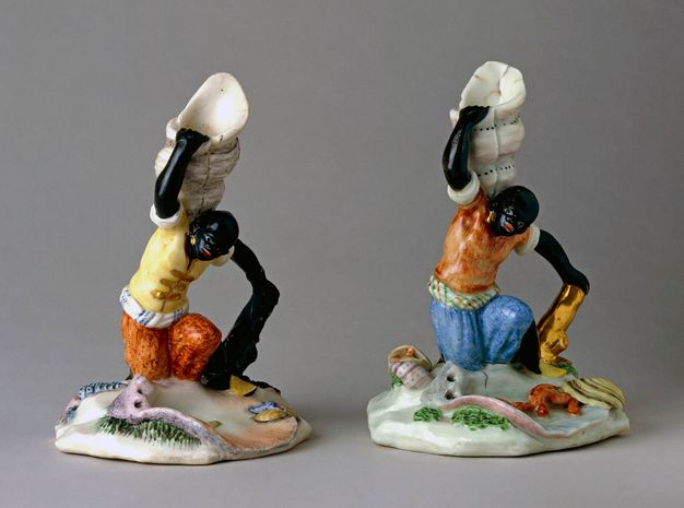 Porcelain. Two candlestick holders are in the shape of people with black-coloured skin kneeling and holding up seashells. The eyes are painted white, the mouths red, and each has a gold earring in the ear we can see.