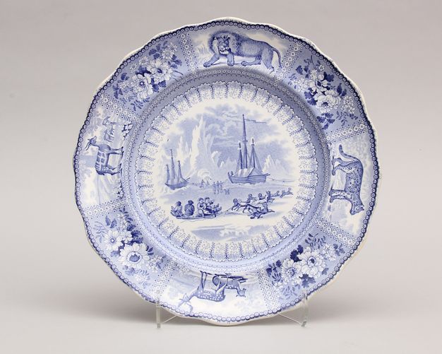 Ceramic. Plate with a gently scalloped edge is illustrated in its centre with two masted ships and people pulled on a sled in tones of pale blue. Animals and floral designs alternate around the rim.