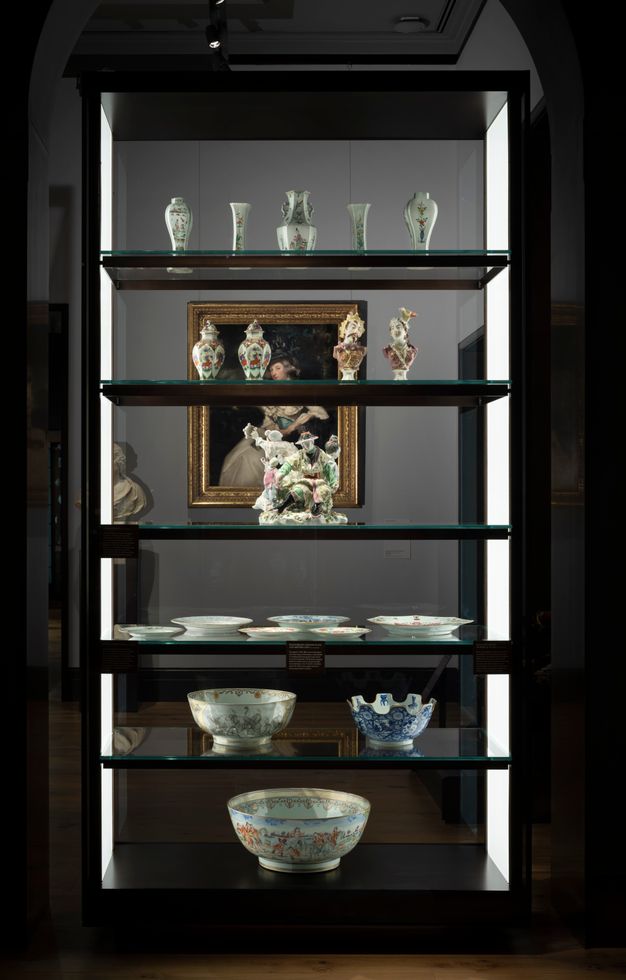 Installation. Six shelves hold porcelains in a case with glass on both long sides so we look through it. From the bottom up, the first two shelves hold bowls, and then a shelf has several plates. A sculptural piece is next, below two shelves of vases and urns.