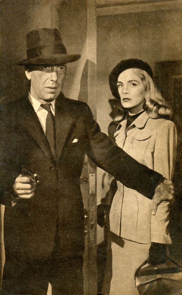 Film still. Black and white image of a pale-skinned man and woman standing near an open door. The man wears a suit and fedora, and holds up revolver in one hand. His other hand reaches across the woman’s body and clutches her far arm. She has long, shiny blond hair under a black cap, and wears a skirt suit and black gloves.