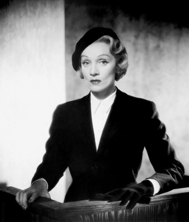 Film still. A pale-skinned woman with curly blond hair arranged carefully under a black beret rests both hands on a witness stand. She wears a tailored black jacket and white shirt, one black glove, and the other hand is bare. Her high cheekbones cast shadows in the bright light.