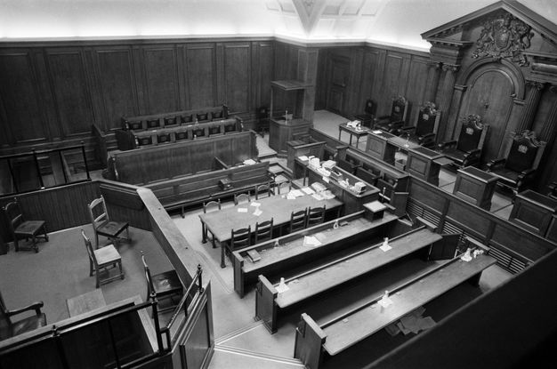 Black and white photograph. We look down onto long tables and rows of chairs in a courtroom. A few papers and water jugs are scattered across and beneath the tables.