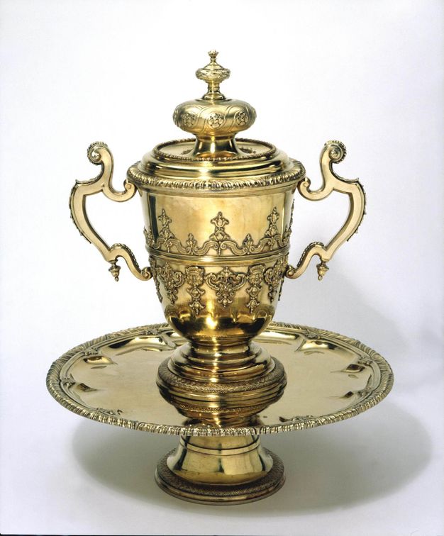 Metalwork. A lidded cup sits on a circular, footed tray. All three pieces are coloured gold. The cup has two scroll handles, and the body of the cup is decorated with strapwork and ornamental designs in relief. The lid has a bulbous and then smaller knob beneath a delicate finial.