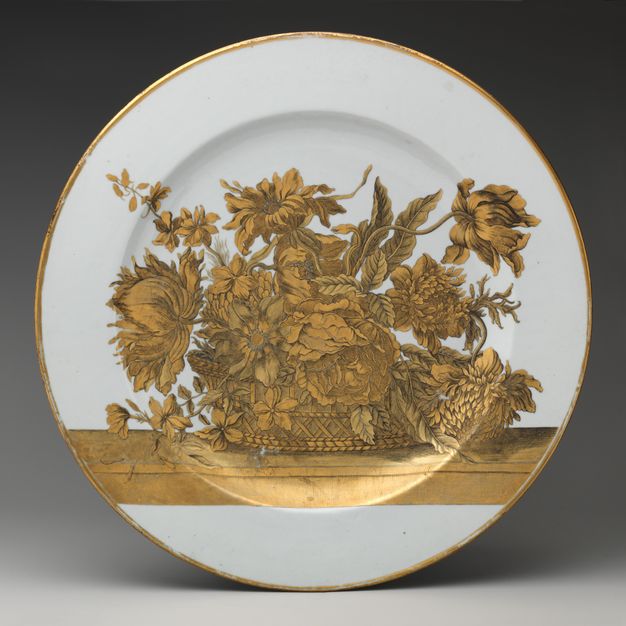 Porcelain. A white plate is decorated with a basket of flowers on a ledge, all in gold.