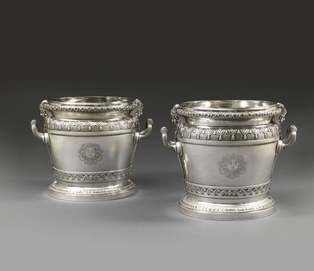Metalwork. Two silver wine coolers have ornamental moulding and designs around the feet, the bottom of the body, the shoulders, and mouth of the flaring rim. Upwards curving handles are on either side of each tub. A coat of arms is incised on the front centre of each cooler.