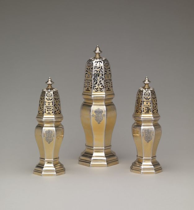 Metalwork. Three different sized casters would have been used like salt and pepper shakers but for sugar, dry mustard, and pepper. Each has an elongated top pierced with scrolls and floral designs. The bodies are six-sided, and each is incised with a coat of arms and the letters “GR”. Some of the gold surface has worn away to show the silver beneath.