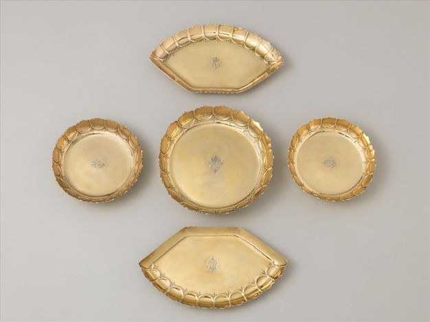 Metalwork. Five gold dishes with fluted, scalloped edges are stamped with a coat of arms and the letters “GR”. Three are round, and one is larger than the other two. The final two are flat on one side and curve in a segment of an arch on the opposite side.