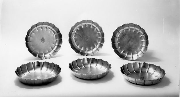 Metalwork. Six silver dishes with fluted and scalloped rims. A coat of arms is stamped into the centre of each, and is flanked by the letters “AR”.