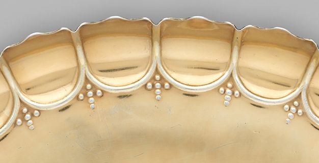 Metalwork. Detail view of a scalloped, fluted edge of a gold-coloured dish. Each scallop is ridged along its inner edge, and an ornament made up of six dots creating a cross is tucked into the space where the curves meet.