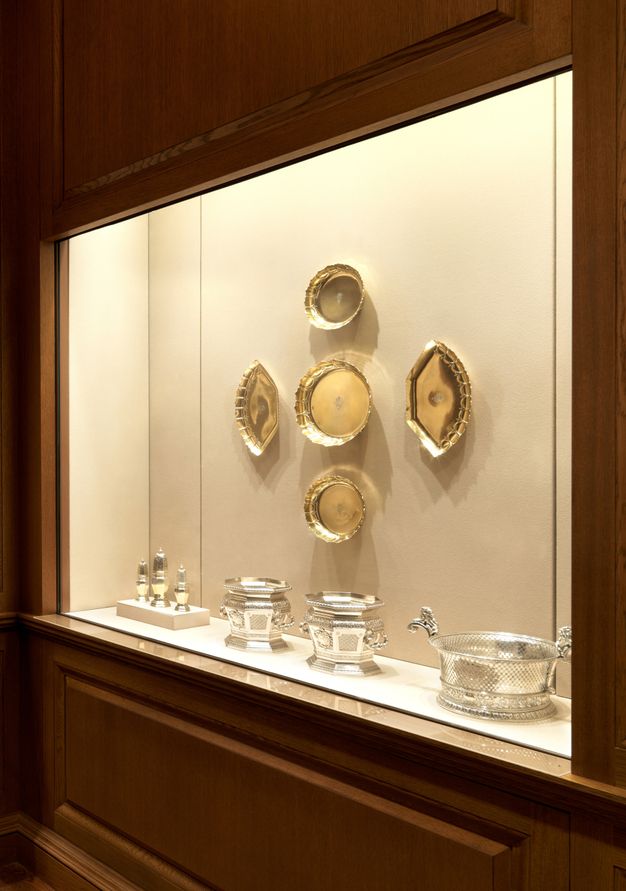 Installation. Silver and gold-coloured vessels and plates are displayed in a case built into a wall. Five gold circular or semi-circular plates are arranged on the wall. A set of three silver casters, a pair of silver wine coolers, and a silver tub line the ledge below.